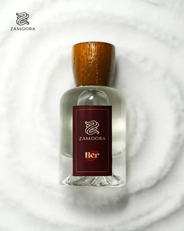 Zamoora HER perfume bottle for women placed among pink flowers with a purple background, highlighting fruity top notes of strawberry, raspberry, blackberry, sour cherry, black currant, mandarin orange and lemon, a heart note of violet jasmine, and base notes of musk, vanilla, cashmeran, woody notes, amber, oakmoss and patchoulI.